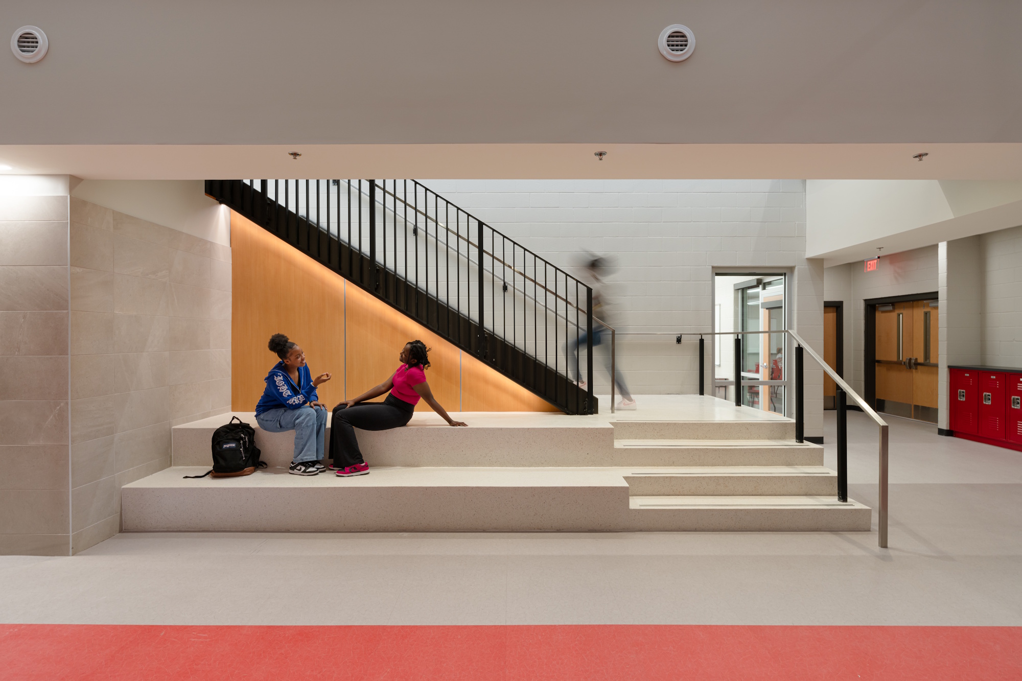 Two people seated on a long bench in front of a staircase going up to the next floor.